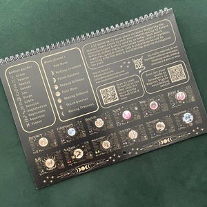 May include: A spiral-bound, black 12-month moon phase calendar. It features zodiac signs, moon phases, and associated tarot cards. The calendar includes text such as "Moon Phases" and "Zodiac Guide". The calendar is designed to aid in working with the moon and manifesting desires.