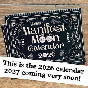 May include: A black spiral-bound wall calendar titled Doomcat's Manifest with the Moon Calendar 2026. The cover features gold celestial illustrations, including moons and stars, with the text This is the 2026 calendar 2027 coming very soon!