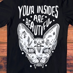 May include: Black t-shirt with a white graphic of a sphinx cat with a pentagram on its forehead. The text "YOUR INSIDES ARE BEAUTIFUL" is above the cat.