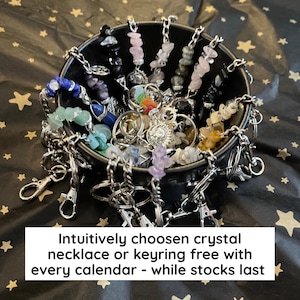 May include: A black bowl filled with various crystal keyrings and necklaces. The crystals are in a variety of colors, including blue, green, pink, and yellow. The keyrings have silver clasps. Text on the image reads: "Intuitively chosen crystal necklace or keyring free with every calendar - while stocks last."