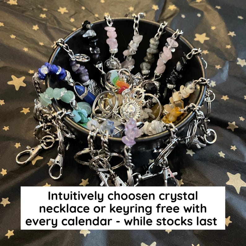 May include: A black bowl filled with various crystal keyrings and necklaces. The crystals are in a variety of colors, including blue, green, pink, and yellow. The keyrings have silver clasps. Text on the image reads: "Intuitively chosen crystal necklace or keyring free with every calendar - while stocks last."