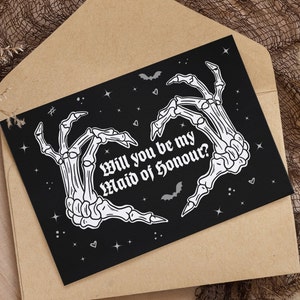 May include: Black card with white skeleton hands forming a heart shape. The text reads "Will you be my Maid of Honour?" with bats and stars in the background.