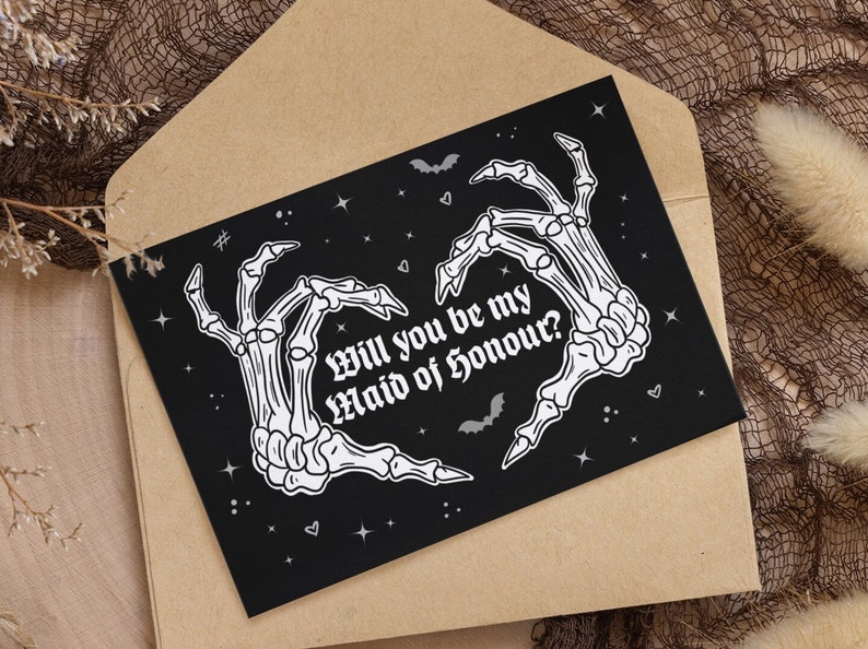 May include: Black card with white skeleton hands forming a heart shape. The text reads "Will you be my Maid of Honour?" with bats and stars in the background.