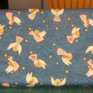 Könnte beinhalten: Blauer Stoff mit einem sich wiederholenden Muster aus weißen Engeln mit rosa und roten Akzenten. Die Engel sind von kleinen gelben Sternen und weißen Blumenmustern umgeben.