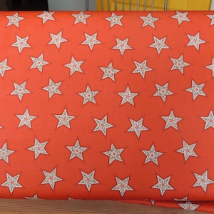 May include: A roll of orange fabric with a repeating pattern of white stars. Each star has a detailed floral design in the center. The fabric is likely used for crafting or sewing projects.