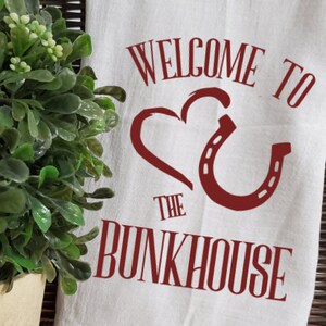May include: White kitchen towel with red text that says "Welcome to the Bunkhouse" with a red heart and horseshoe design.