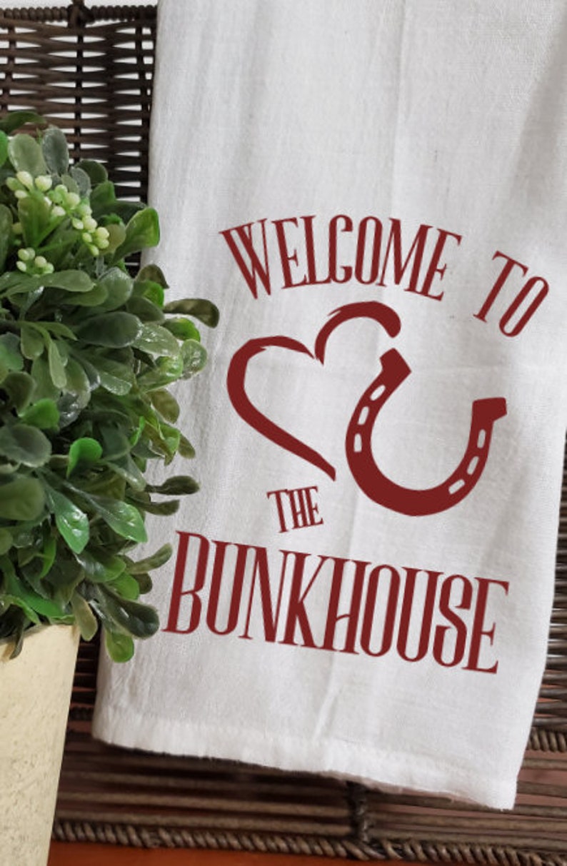 May include: White kitchen towel with red text that says "Welcome to the Bunkhouse" with a red heart and horseshoe design.