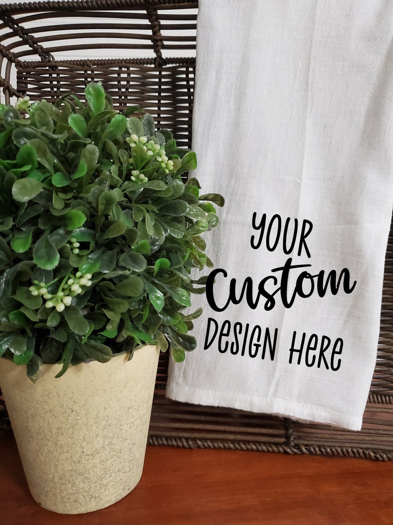 May include: White kitchen towel with the text "YOUR Custom Design Here" in black lettering. The towel is folded and displayed on a wicker basket with a potted green plant.