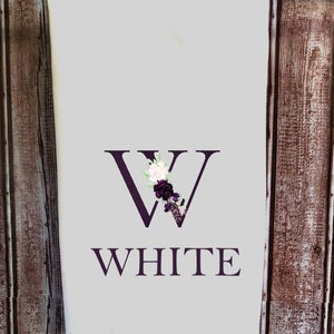 May include: White kitchen towel with a purple floral design and the letter "W" in the center. The word "WHITE" is printed below the letter.