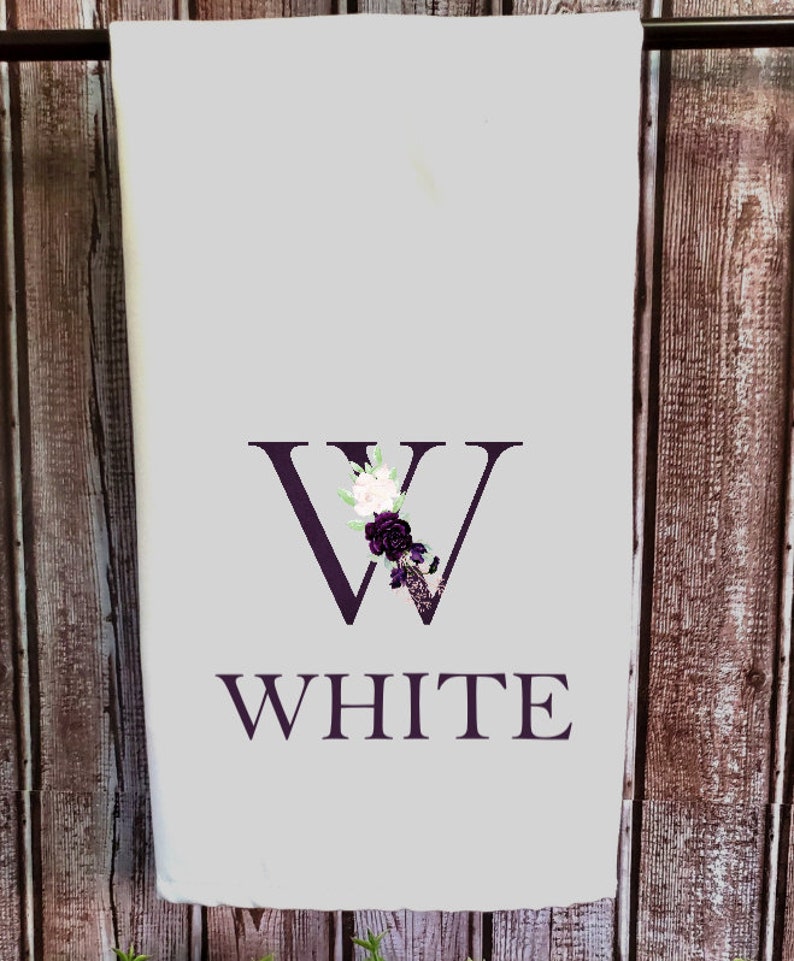 May include: White kitchen towel with a purple floral design and the letter "W" in the center. The word "WHITE" is printed below the letter.