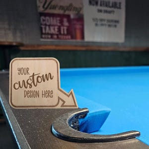 May include: A wooden sign with the text "YOUR custom DESIGN HERE" in a stylized font. The sign has a rounded square shape with an arrow pointing right. The sign is placed on the edge of a pool table with a blue felt surface.