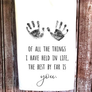 May include: A white kitchen towel with two black handprints and the text "OF ALL THE THINGS I HAVE HELD IN LIFE, THE BEST BY FAR IS you."