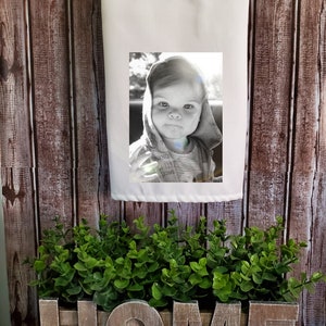 May include: A white kitchen towel with a black and white photo of a young child wearing a hooded sweatshirt. The towel is hanging on a wooden wall with the word "HOME" spelled out in wooden letters in front of it.