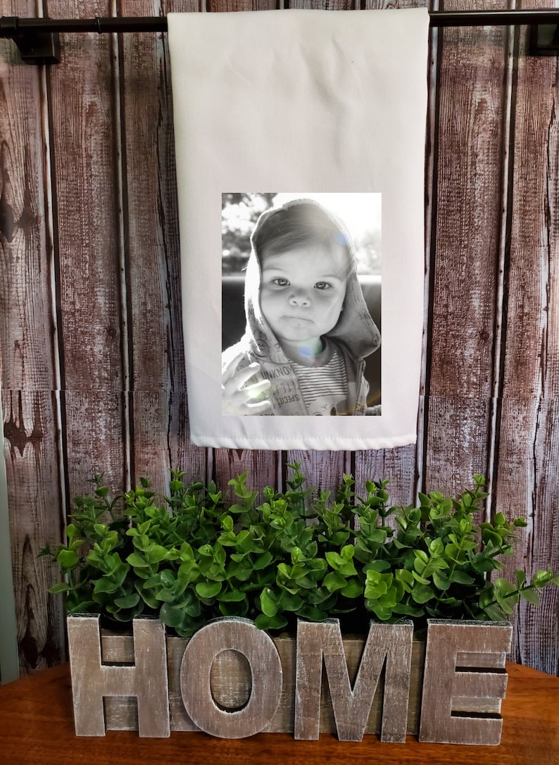 May include: A white kitchen towel with a black and white photo of a young child wearing a hooded sweatshirt. The towel is hanging on a wooden wall with the word "HOME" spelled out in wooden letters in front of it.