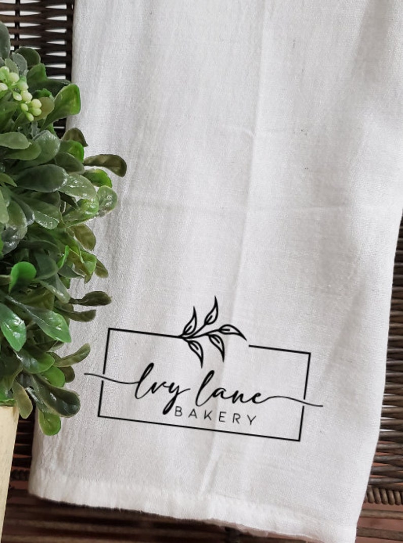 May include: White kitchen towel with a black logo that says "ivy lane bakery" with a simple leaf design.