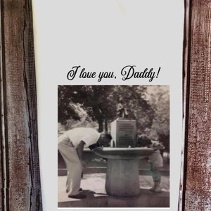 May include: A black and white photograph of a father and child drinking from a water fountain. The photo is printed on a white tea towel with the text "I love you, Daddy!" printed above the photo.