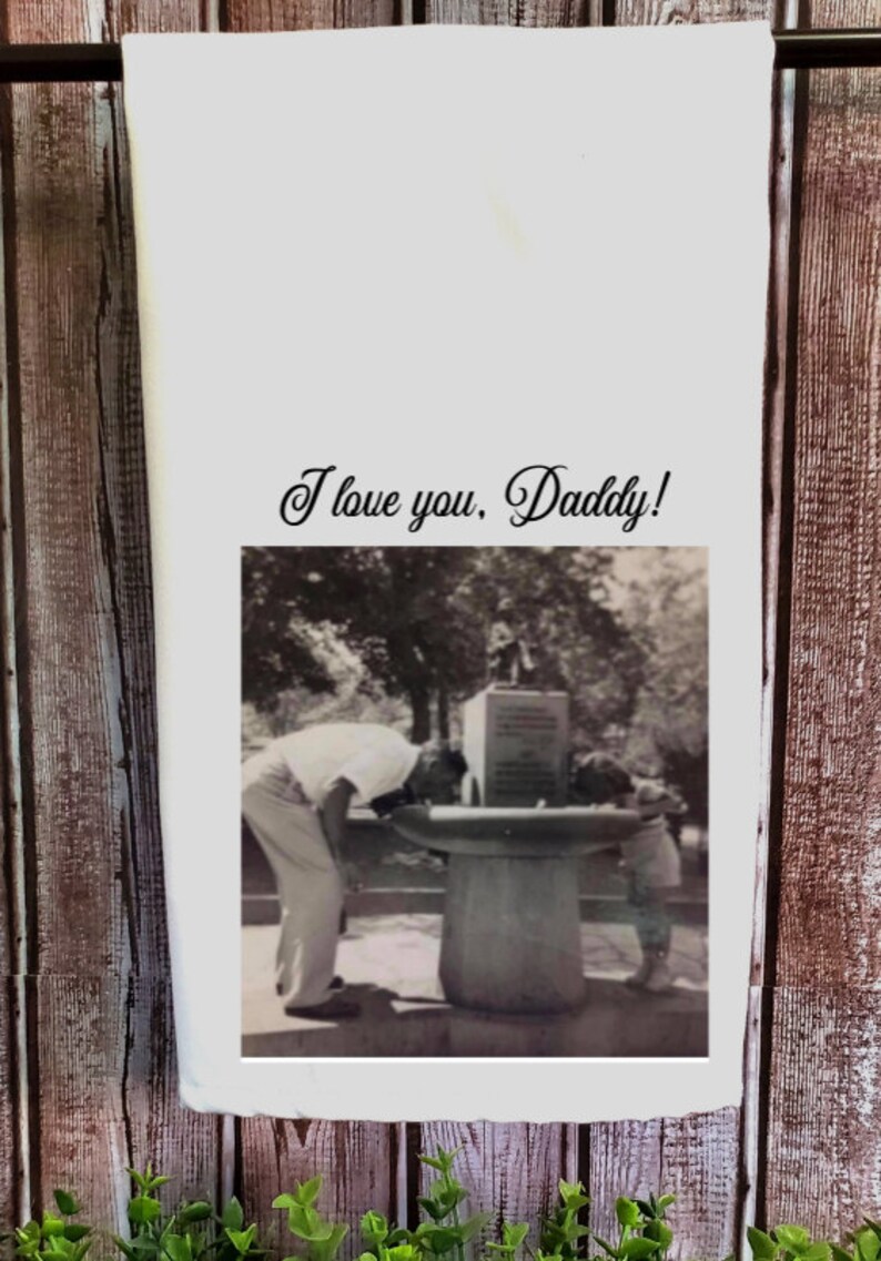 May include: A black and white photograph of a father and child drinking from a water fountain. The photo is printed on a white tea towel with the text "I love you, Daddy!" printed above the photo.
