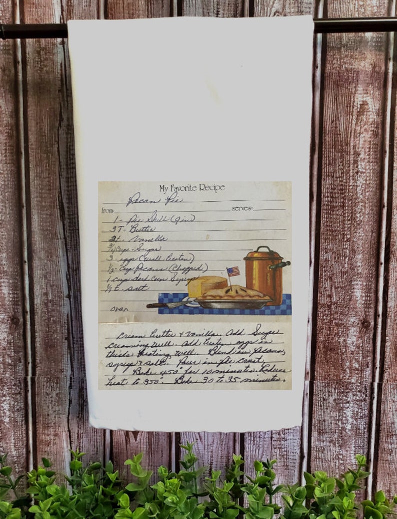 May include: A white tea towel with a vintage recipe card printed on it. The recipe card is for pecan pie and includes a list of ingredients and instructions. The card is illustrated with a pie and a copper pot.
