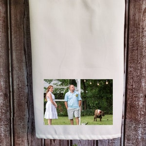 May include: A white kitchen towel with a photo of a couple standing in a grassy area with a dog in the background. The photo is printed on the towel.