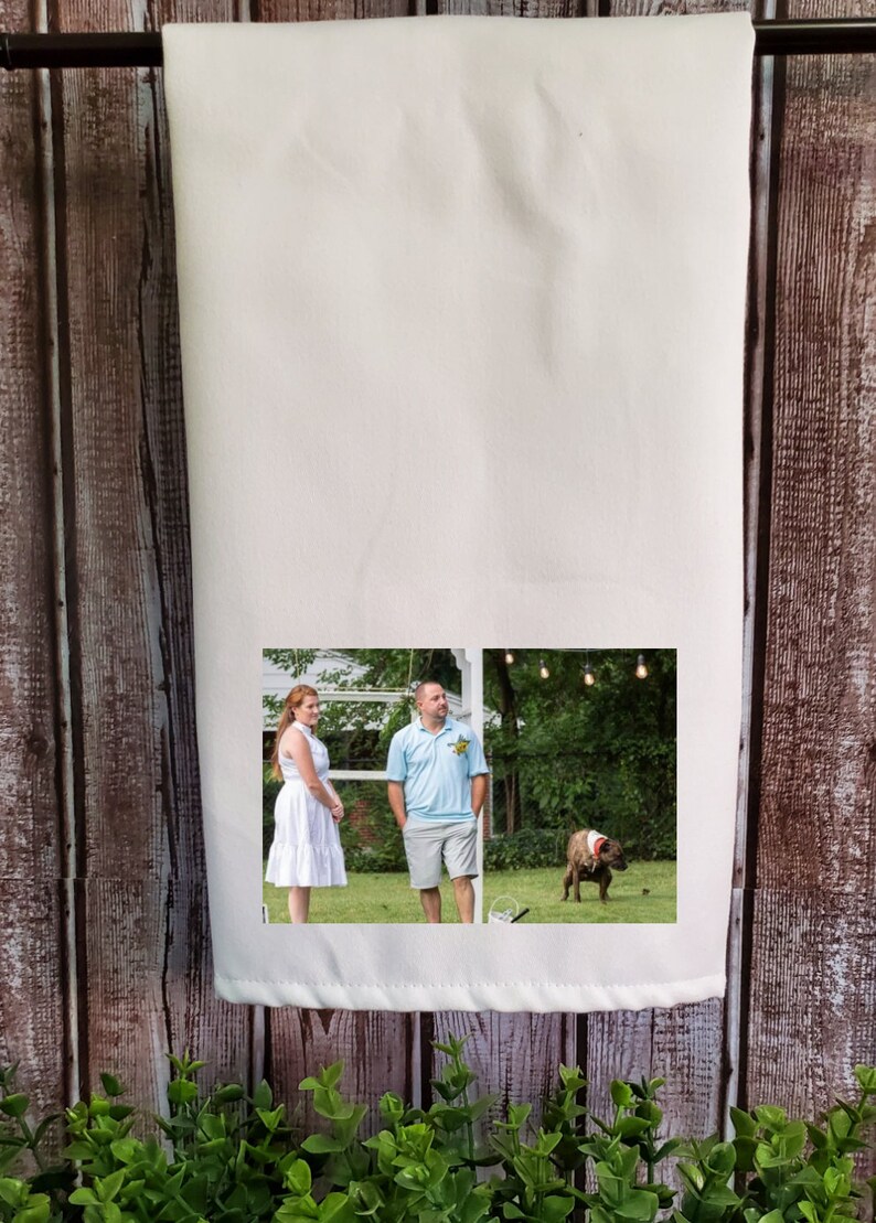 May include: A white kitchen towel with a photo of a couple standing in a grassy area with a dog in the background. The photo is printed on the towel.
