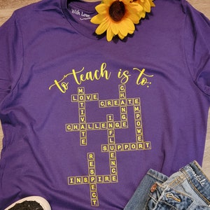 May include: Purple t-shirt with the words "to teach is to." in yellow lettering. The words are arranged in a crossword puzzle format with words like "love", "create", "challenge", "inspire", and "support".