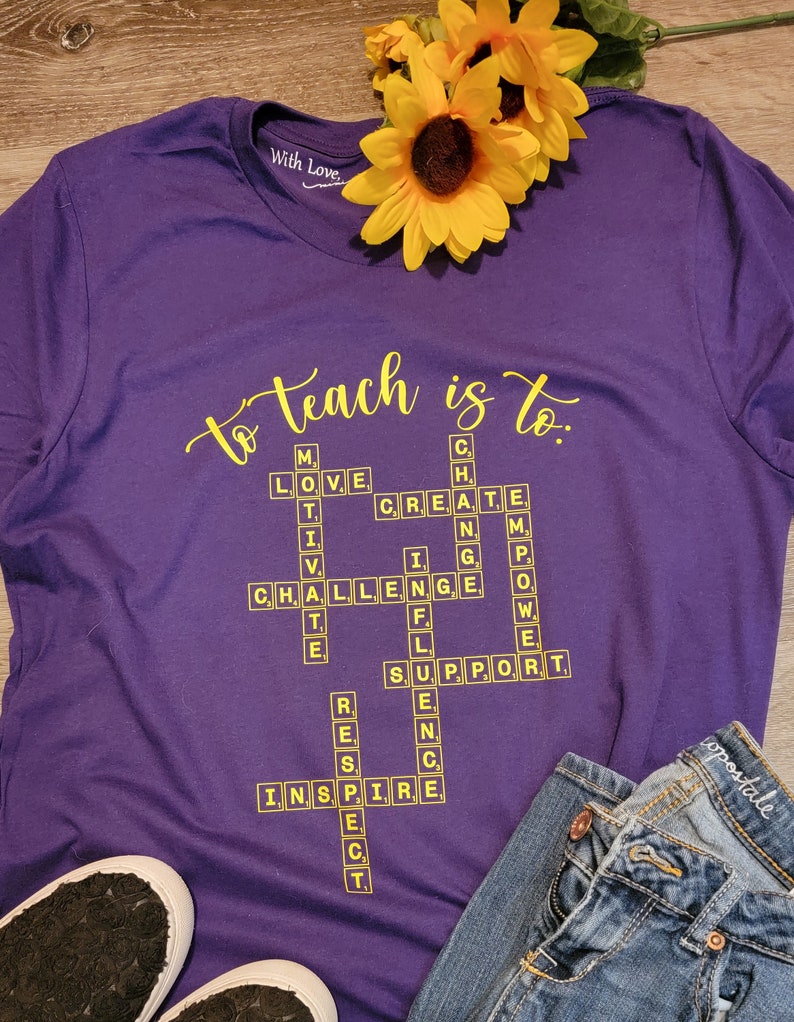 May include: Purple t-shirt with the words "to teach is to." in yellow lettering. The words are arranged in a crossword puzzle format with words like "love", "create", "challenge", "inspire", and "support".