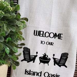 May include: White kitchen towel with black text and graphics. The text reads "Welcome to our Island Oasis". The graphics include two chairs, a fire pit, and two glasses.