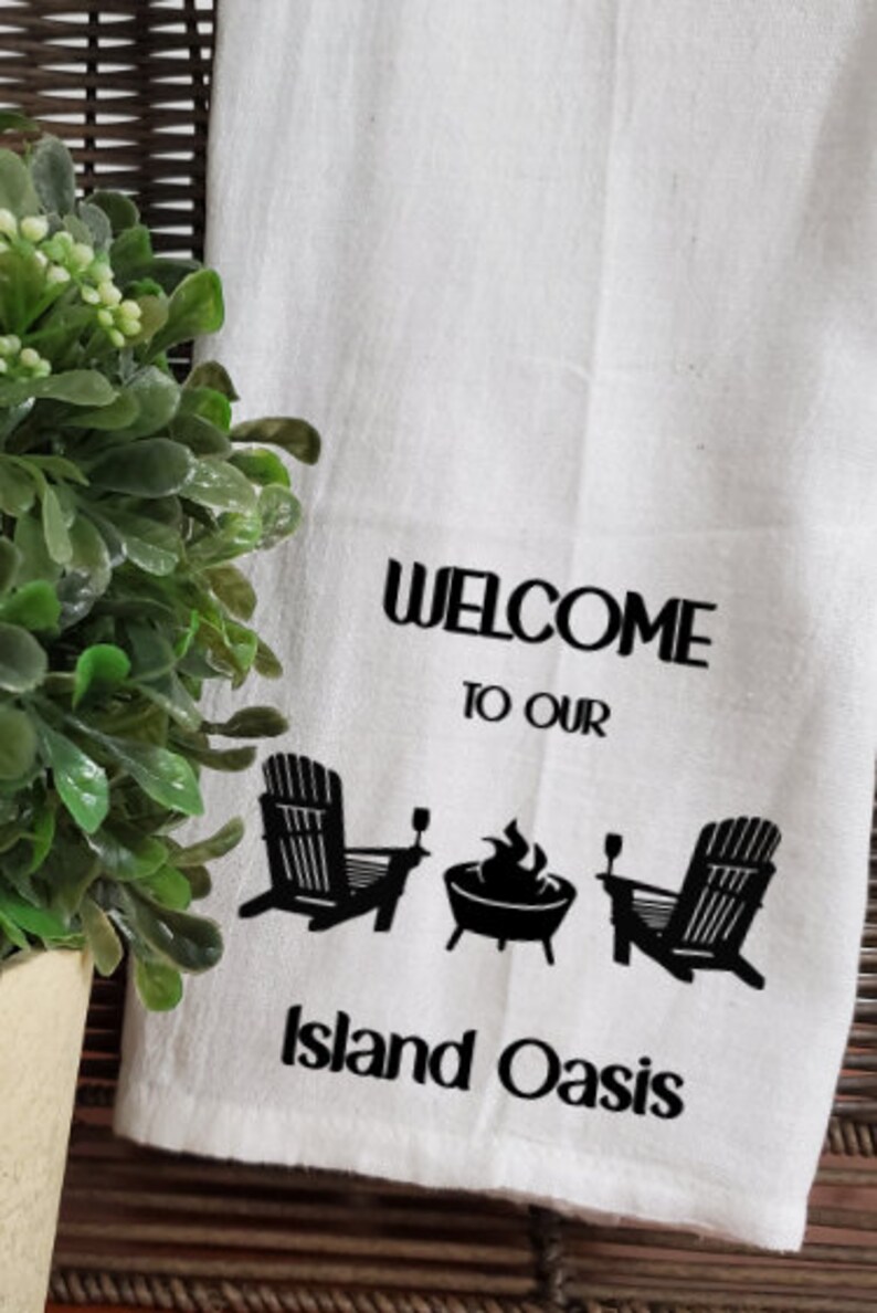 May include: White kitchen towel with black text and graphics. The text reads "Welcome to our Island Oasis". The graphics include two chairs, a fire pit, and two glasses.