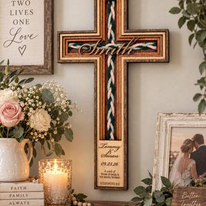 May include: A wooden wall cross featuring a braided cord of teal, pink, and dark green strands. The cross is engraved with the name Smith, the names Tommy & Susan, the date 09.25.26, and the text A cord of three strands is not easily broken.