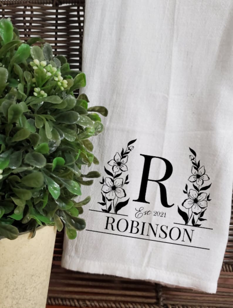 May include: White kitchen towel with a black floral design and the letter "R" in the center. The text "Est. 2021 ROBINSON" is printed below the letter.