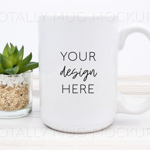 May include: A white ceramic mug with a large handle, featuring the text "YOUR design HERE" in black script. A small succulent plant in a glass container is to the left of the mug. The mug is on a light wood surface.