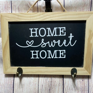 May include: A wooden framed chalkboard sign with the text "Home Sweet Home" in white lettering. The sign has two hooks on the bottom.