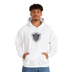 May include: White hooded sweatshirt with a gray shield graphic on the front. The graphic features a stylized wing design and text. The sweatshirt has a kangaroo pocket and a drawstring hood. The model is wearing blue jeans.