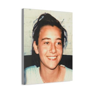 Könnte beinhalten: Leinwanddruck mit einer lächelnden Person mit dunklem Haar. Die Person trägt ein hellblaues Hemd und eine Halskette. Der Hintergrund ist eine hellfarbige Wand mit einem dezenten Muster. Das Bild ist ein Porträt.