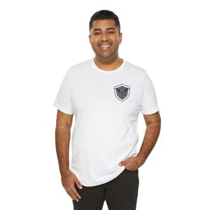 Op de afbeelding: Wit T-shirt met een klein, grijs schildembleem op de linkerborst. Het embleem heeft een gestileerd ontwerp in een schildvorm. Het shirt is een klassieke ronde hals.