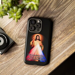 May include: Black phone case featuring an image of Jesus with a halo, radiating light. The image includes the text "His mercies never come to an end (Lamentations 3:22)". The case is on a wooden surface.