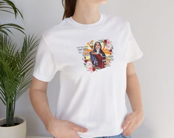 Op de afbeelding: Wit t-shirt met een afbeelding van een religieuze figuur die een gouden voorwerp vasthoudt, omringd door bloemen en tekst. De tekst luidt "Let the love you have in your heart..." en "...shine outwardly in your deeds."