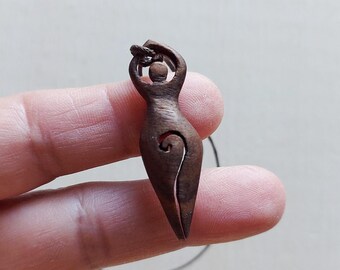 May include: A dark brown wooden pendant carved in the shape of a stylized female figure. The pendant features a spiral design on the torso and is suspended from a thin, dark cord. The figure's arms are raised above its head.
