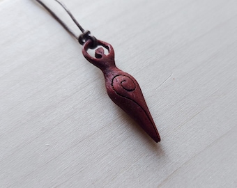 May include: A maroon wooden pendant necklace. The pendant is carved in the shape of a female figure with a spiral design. It hangs from a dark brown cord, giving a natural, handcrafted look.