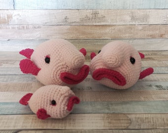 May include: Three crocheted plush toys shaped like blobfish. The largest blobfish is pink with a red mouth and black eyes. The medium blobfish is pink with a red mouth and black eyes. The smallest blobfish is pink with a red mouth and black eyes.