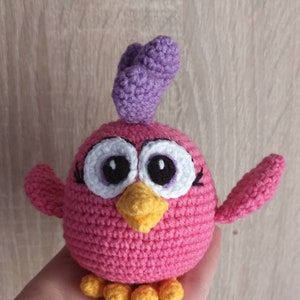May include: A pink crocheted bird with a purple crest, large white eyes with black pupils, and a yellow beak. The bird has yellow feet and is holding its wings out.