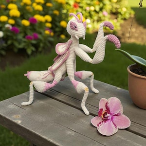May include: A handmade crochet praying mantis in white and pink, posed on a wooden surface. The insect has detailed features, including large eyes and articulated limbs. A pink and purple orchid flower lies nearby, with a blurred garden background.