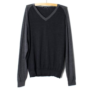 Peut inclure: Un pull à col en V et manches longues avec un torse gris anthracite foncé et des manches gris moyen contrastantes. Le vêtement est présenté sur un cintre en bois et comprend un col, des poignets et un ourlet côtelés, avec une étiquette visible.