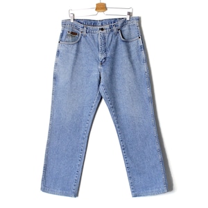 Op de afbeelding: Lichtblauwe spijkerbroek hangend aan een houten hanger. De jeans heeft een klassiek ontwerp met vijf zakken, een knoop- en ritssluiting en een kleine bruine patch op de voorzak. De jeans is licht gewassen en lijkt vintage.