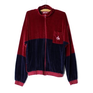 Puede incluir: Una chaqueta de terciopelo rojo y azul con cremallera y un pequeño logotipo bordado en el bolsillo.