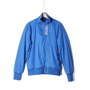 Könnte beinhalten: Eine blaue Reißverschlussjacke mit geripptem Kragen und Bündchen. Die Jacke hat den Schriftzug "Bench" in Silber auf der Vorderseite.