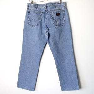 Op de afbeelding: Lichtblauwe spijkerbroek hangend aan een houten hanger. De jeans heeft een klassiek ontwerp met vijf zakken, met de Wrangler-logopatch op de achterzak. De jeans heeft een rechte pijp.
