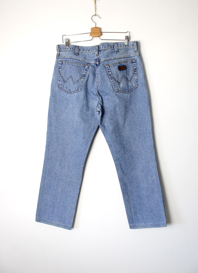 Op de afbeelding: Lichtblauwe spijkerbroek hangend aan een houten hanger. De jeans heeft een klassiek ontwerp met vijf zakken, met de Wrangler-logopatch op de achterzak. De jeans heeft een rechte pijp.
