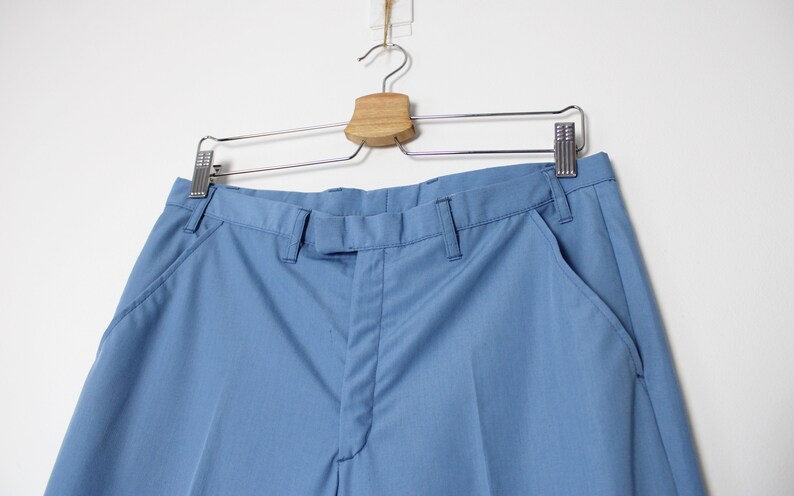 K&ouml;nnte beinhalten: Eine hellblaue Hose, die an einem Holzb&uuml;gel h&auml;ngt. Die Hose hat einen Rei&szlig;verschluss und einen Knopf vorne, zwei Vordertaschen und zwei Ges&auml;&szlig;taschen.