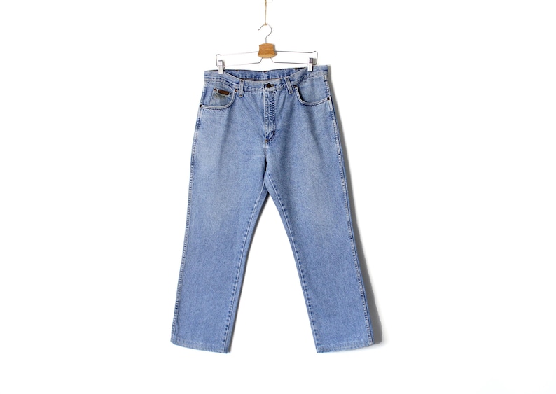 Op de afbeelding: Lichtblauwe spijkerbroek hangend aan een houten hanger. De jeans heeft een klassiek ontwerp met vijf zakken, een knoop- en ritssluiting en een kleine bruine patch op de voorzak. De jeans is licht gewassen en lijkt vintage.